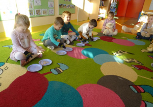 Czwórka dzieci z założonymi rękawiczkami przekłada wykałaczki z jednego talerzyka na drugi. Reszta dzieci obserwuje z zaciekawieniem.